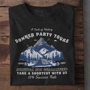 Może przedstawiać: Czarna koszulka z graficznym wzorem przedstawiającym góry, drzewa i namiot. Tekst głosi "Donner Party Tours" i "Survival Not Guaranteed". Koszulka zawiera również lata 1846 i 1847 oraz frazę "55% Success Rate."