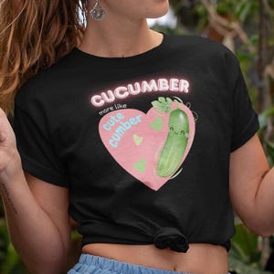 May include: Black t-shirt with a pink heart-shaped graphic featuring a smiling cucumber and the text "Cucumber more like cute cumber." The person is wearing a blue skirt and holding a white mug. The background is filled with green plants.