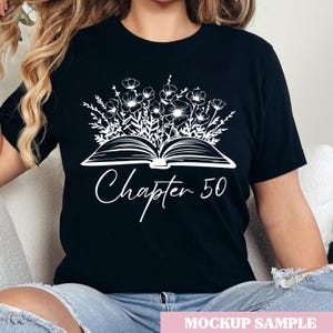Puede incluir: Camiseta negra con un diseño floral blanco que emerge de un libro abierto. El texto "Chapter 50" está escrito en cursiva debajo del libro. La camiseta es una muestra de maqueta.