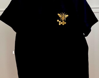 Może przedstawiać: Czarny t-shirt ze złotym wzorem pszczoły i plastra miodu na górnej lewej piersi. Pszczoła jest szczegółowa ze skrzydłami i segmentami ciała. Koszulka wisi na drewnianym wieszaku.