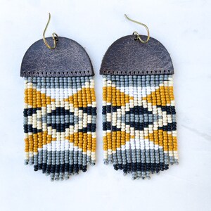 May include: A pair of beaded fringe earrings with a brown leather backing. The earrings feature a geometric pattern of black, white, and mustard yellow beads.