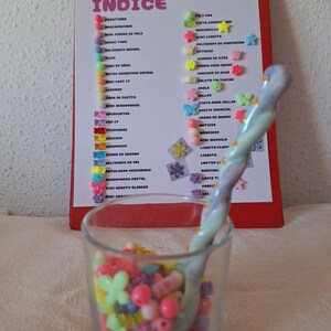 Puede incluir: Un vaso transparente lleno de cuentas de colores y una cuchara decorativa. Un portapapeles rojo con una lista de artículos en español está en el fondo, sujeto por un clip dorado. La lista se titula "LUCKY BOX" e "ÍNDICE".