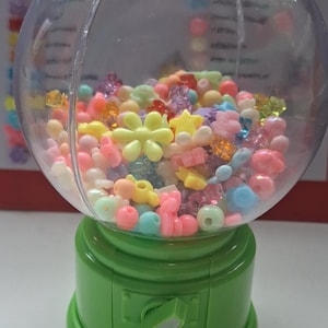 Puede incluir: Una máquina de chicles verde llena de cuentas de colores en varias formas, incluyendo flores y estrellas. El globo de plástico transparente contiene una variedad de cuentas de colores pastel. La máquina tiene una base verde y una ranura de dispensación.
