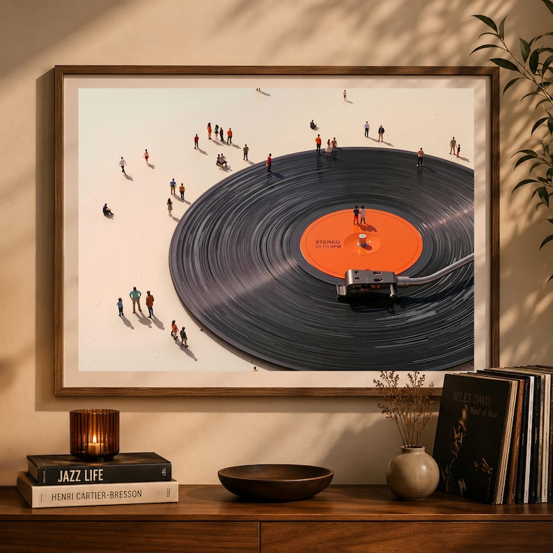 May include: A framed art print featuring a large vinyl record with tiny figures scattered across its surface and the surrounding area. The record has an orange label with text. The artwork is displayed on a wooden cabinet with books, a candle, and a small vase.