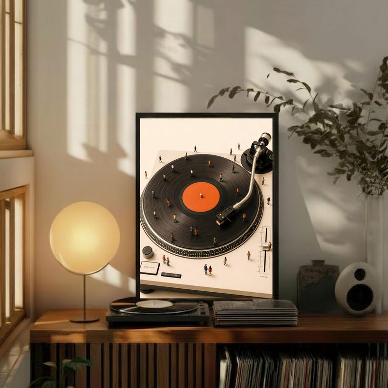 May include: A framed art print featuring a high-angle view of a white turntable with a black vinyl record. The record has a bright orange center label. Tiny miniature figures are scattered across the surface of the record and the turntable base.