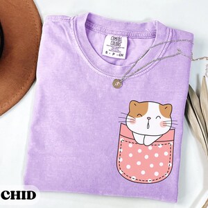May include: A light purple short-sleeved t-shirt with a crew neckline. The shirt features a graphic of a white and brown cat peeking out of a pink pocket with white polka dots. The interior neck label reads Comfort Colors, S-P-CH, and 100% cotton.
