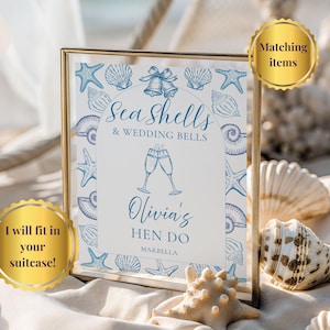 May include: A framed sign with a gold border, featuring blue seashell and starfish illustrations. The text reads "Sea Shells & Wedding Bells" and "Olivia's HEN DO MARBELLA". A gold sticker says "I will fit in your suitcase!"