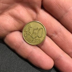 May include: A gold-coloured 50 Euro cent coin. The coin displays the number 50, with the words "EURO CENT" below. The coin is held in a hand.