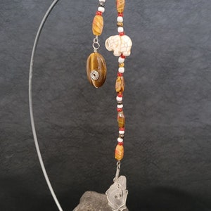 May include: A beaded necklace featuring a tiger's eye stone, a white elephant charm, and a clear quartz crystal wrapped in wire. The necklace is displayed on a metal stand.