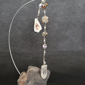 May include: A clear quartz crystal point hangs from a silver wire with colorful beads and a shell pendant. The crystal is attached to a gray rock base.