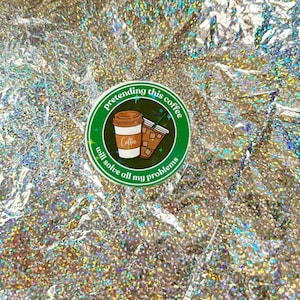 Könnte beinhalten: Ein runder Aufkleber mit grünem Rand und dem Text "pretending this coffee will solve all my problems". Der Aufkleber zeigt Illustrationen von zwei Kaffeetassen. Der Hintergrund ist ein glänzendes, irisierendes Silber.