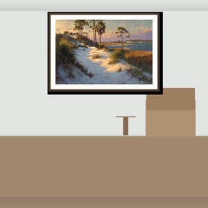 May include: A framed landscape painting shows a coastal scene with a sandy path, dunes, and trees under a sunset sky. The artwork is in a dark brown frame, hanging on a light grey wall.