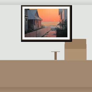 May include: A framed landscape art print showing a coastal scene at sunset. The artwork features a wooden building, a white picket fence, and a path leading to the ocean under an orange sky. The frame is dark brown.