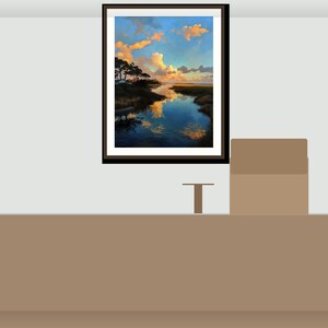 May include: A framed landscape painting shows a tranquil waterscape with a reflective river, silhouetted trees, and a vibrant sky of blue, orange, and yellow hues. The artwork is displayed above a brown counter.