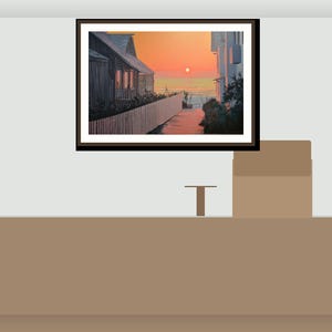 May include: A framed landscape painting showing a coastal scene at sunset. The artwork features a path leading to the sea, with buildings and a white picket fence. The sky is a gradient of orange and yellow.