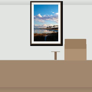 May include: A framed landscape print showing a coastal scene with a blue sky, white clouds, and water reflecting the sky. The artwork is in a dark brown frame and hangs on a light grey wall. A small table and a box are in the foreground.
