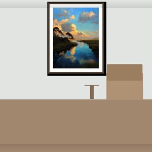 May include: A framed landscape art print showing a tranquil river scene with trees and a cloudy sky. The artwork has a dark brown frame and is displayed on a light grey wall above a brown surface.