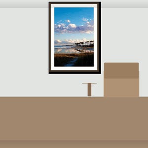 May include: A framed landscape print depicting a coastal scene with a blue sky, white clouds, and a body of water reflecting the sky. The print is in a black frame, hanging above a brown surface.