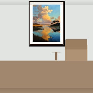 May include: A framed landscape painting shows a tranquil coastal scene with a colourful sky of orange and blue tones. The artwork reflects clouds and trees in the calm water, creating a peaceful atmosphere. The painting is displayed above a brown table.