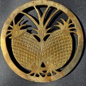 May include: A round, gold-colored metal trivet featuring a detailed pineapple design. The trivet has a circular frame with an openwork design and two pineapples in the center. The pineapple design is raised and textured.