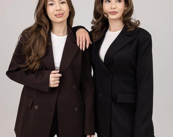 May include: Two women wearing matching suits. One suit is a dark brown color, and the other is black. Both suits have a double-breasted blazer and matching pants. The women are standing close together, with one woman's arm resting on the other's shoulder.