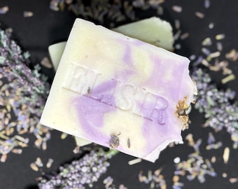 May include: A bar of handmade soap with a lavender and cream swirl design, stamped with the word "ELISIR". The soap is surrounded by dried lavender buds and sprigs, creating a natural and aromatic aesthetic.