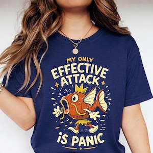 Könnte beinhalten: Marineblaues T-Shirt mit einem Cartoon-Fisch-Design. Der Text auf dem Shirt lautet "MY ONLY EFFECTIVE ATTACK IS PANIC" in Gelb und Weiß. Das Shirt wird mit blauen Jeans und weißen Turnschuhen getragen.