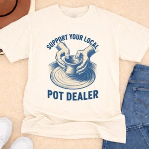 May include: Ivory-colored t-shirt with the text "SUPPORT YOUR LOCAL POT DEALER" in blue. The graphic shows hands shaping a pot on a pottery wheel. The shirt is paired with blue jeans and white sneakers.