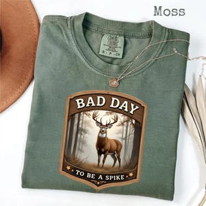 May include: A moss green t-shirt with a graphic featuring a deer in a forest scene. The graphic reads "BAD DAY TO BE A SPIKE" in a wooden-style frame. The shirt has a crew neck and short sleeves. A silver necklace is draped across the shirt.