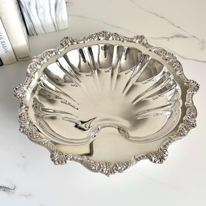 May include: A silver-colored, shell-shaped serving dish with ornate, floral-patterned edges. The dish has a central, fluted design and a smooth, reflective surface. The dish is likely made of metal and is suitable for serving food or as a decorative piece.