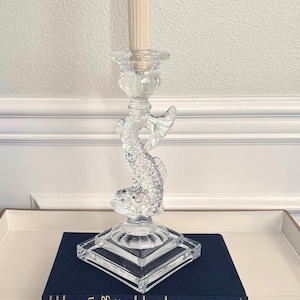 May include: Clear glass candlestick holder featuring a dolphin design. The candlestick has a square base and a detailed dolphin sculpture supporting the candle cup. A cream-colored candle sits in the holder. The candlestick is approximately 10 inches tall.