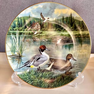 May include: Decorative plate featuring a serene lakeside scene with ducks, a cabin, and lush greenery. The plate has a gold rim and is displayed on a clear stand. The artwork depicts ducks in flight and on the water.