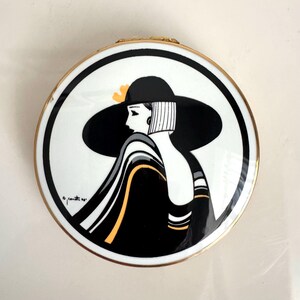 May include: A round, vintage compact with a black and white illustration of a woman in a hat. The woman is wearing a black dress with yellow accents. The compact has a gold-colored rim and a white background.