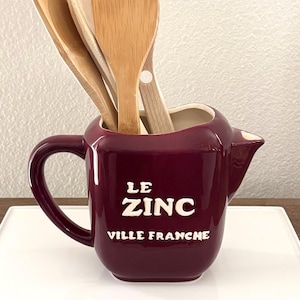 May include: A burgundy ceramic utensil holder with a handle and spout, filled with wooden spoons. The holder has the text "LE ZINC VILLE FRANCHE" in white lettering. The holder is on a white tray.