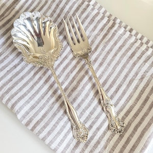 May include: A silver-toned serving spoon and fork set featuring intricate embossed scrollwork and floral detailing on the handles. The spoon has a wide, shell-shaped bowl with scalloped edges, while the fork has three long, slender tines.