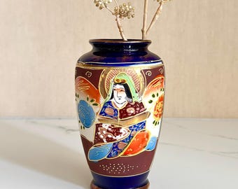 May include: A small ceramic vase featuring a deep blue base and rim with a central illustration of a figure in traditional attire. The design includes gold accents, floral motifs, and a dark red background. A dried branch is placed inside the vase.