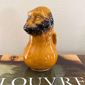 May include: A vintage ceramic pitcher in the shape of a dog's head, with a brown and black glaze. The pitcher has a small handle and a spout. The piece is likely a decorative item or a novelty item.