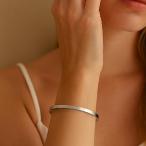 Może przedstawiać: Srebrna bransoletka typu mankiet o minimalistycznym designie. Bransoletka to prosty, zakrzywiony pasek o szerokości około 1,3 cm. Zdjęcie przedstawia bransoletkę na nadgarstku osoby, z neutralnym tłem.