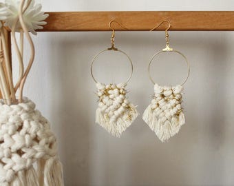 May include: A pair of handmade macrame earrings. Each earring features a gold-colored hoop with a woven cream-colored macrame design and gold accents. The earrings are displayed against a light background.