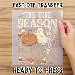 May include: A rectangular DTF transfer sheet featuring the text 'TIS THE SEASON' in a cream font. The design includes a large orange pumpkin, a white ghost, a skeleton, a tombstone marked 'RIP', potion bottles, a haunted house, and candy corn.