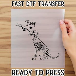 May include: A black and white DTF transfer featuring a skeleton dog being touched by a skeleton hand. The word "Boop" is above the dog's head, with a heart symbol. The text "FAST DTF TRANSFER" and "READY TO PRESS" are also present.