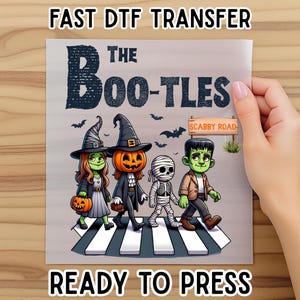 May include: A DTF transfer featuring a Halloween-themed design with the text "The Boo-tles" and "Scabby Road". The image shows cartoon characters dressed as a witch, a pumpkin, a mummy, and Frankenstein crossing a crosswalk. The text "Fast DTF Transfer" and "Ready to Press" are also included.
