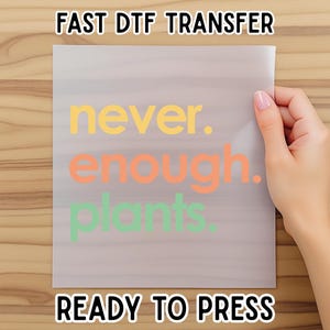 May include: A clear, rectangular DTF transfer sheet with the words "never. enough. plants." in yellow, orange, and green. The sheet is held by a hand and has the text "FAST DTF TRANSFER" and "READY TO PRESS" above and below the design.