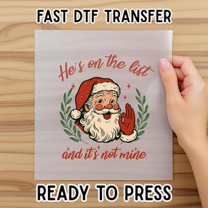 May include: A ready-to-press DTF transfer featuring a vintage Santa Claus design. The image includes the text "He's on the list and it's not mine." Santa is wearing a red hat and gloves, with a white beard and a green wreath.