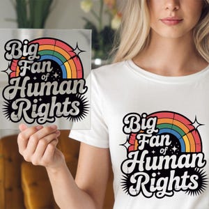 May include: White t-shirt with the text "Big Fan of Human Rights" in a retro design featuring a rainbow and stars. The same design is also on a clear square held in the hand.