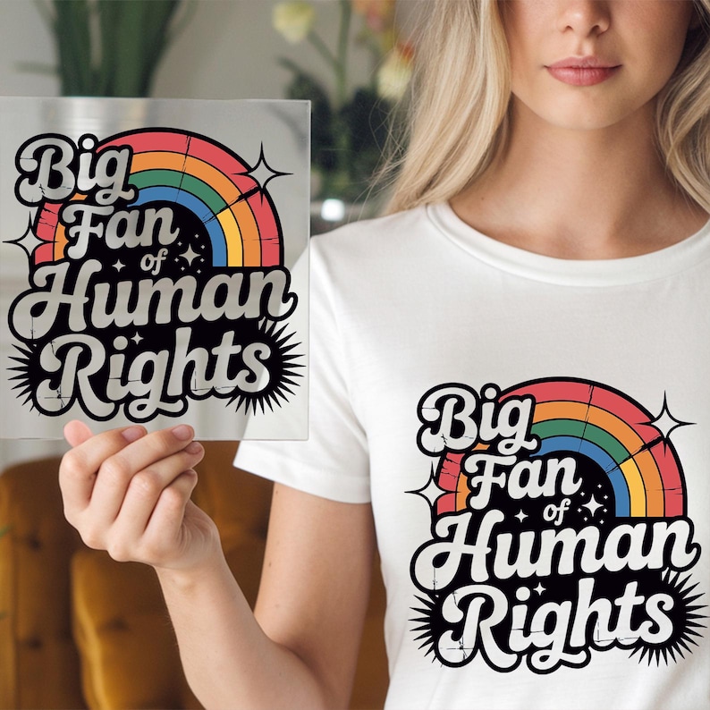 May include: White t-shirt with the text "Big Fan of Human Rights" in a retro design featuring a rainbow and stars. The same design is also on a clear square held in the hand.