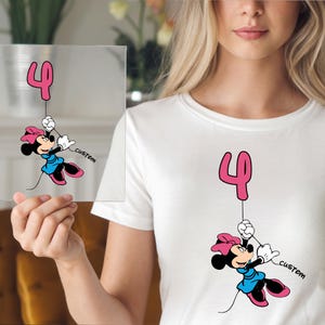 May include: White t-shirt featuring a cartoon Minnie Mouse holding a pink number 4 balloon. Minnie Mouse is wearing a blue dress, pink bow, and black shoes. The word "custom" is printed on the shirt.