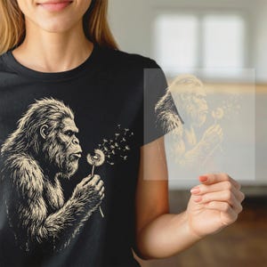 May include: A black t-shirt featuring a graphic illustration of a hairy primate blowing on a dandelion, with seeds drifting into the air. The design is rendered in a light beige, ink-style sketch, showcasing detailed fur texture and facial features.