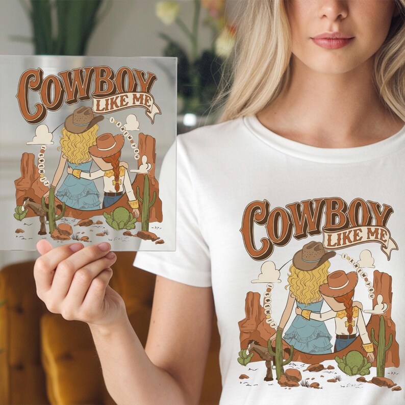 May include: A white t-shirt featuring a graphic illustration of two figures in cowboy hats standing in a desert landscape with cacti and rock formations. The text COWBOY LIKE ME is printed in brown above the figures, with additional text circling them.