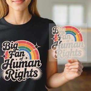 May include: Black t-shirt with a rainbow graphic and the text "Big Fan of Human Rights." The design includes a rainbow, stars, and a sunburst effect. The shirt is made of a soft material.
